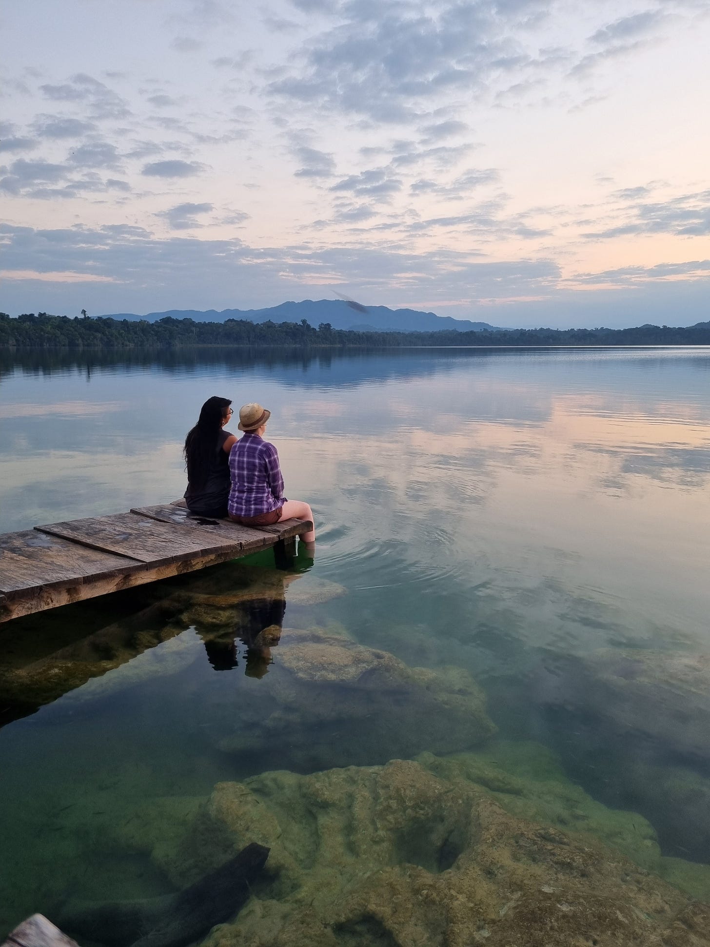 two people sit on a dock over looking a gorgeous lake filled with blues and pinks reflecting the sky