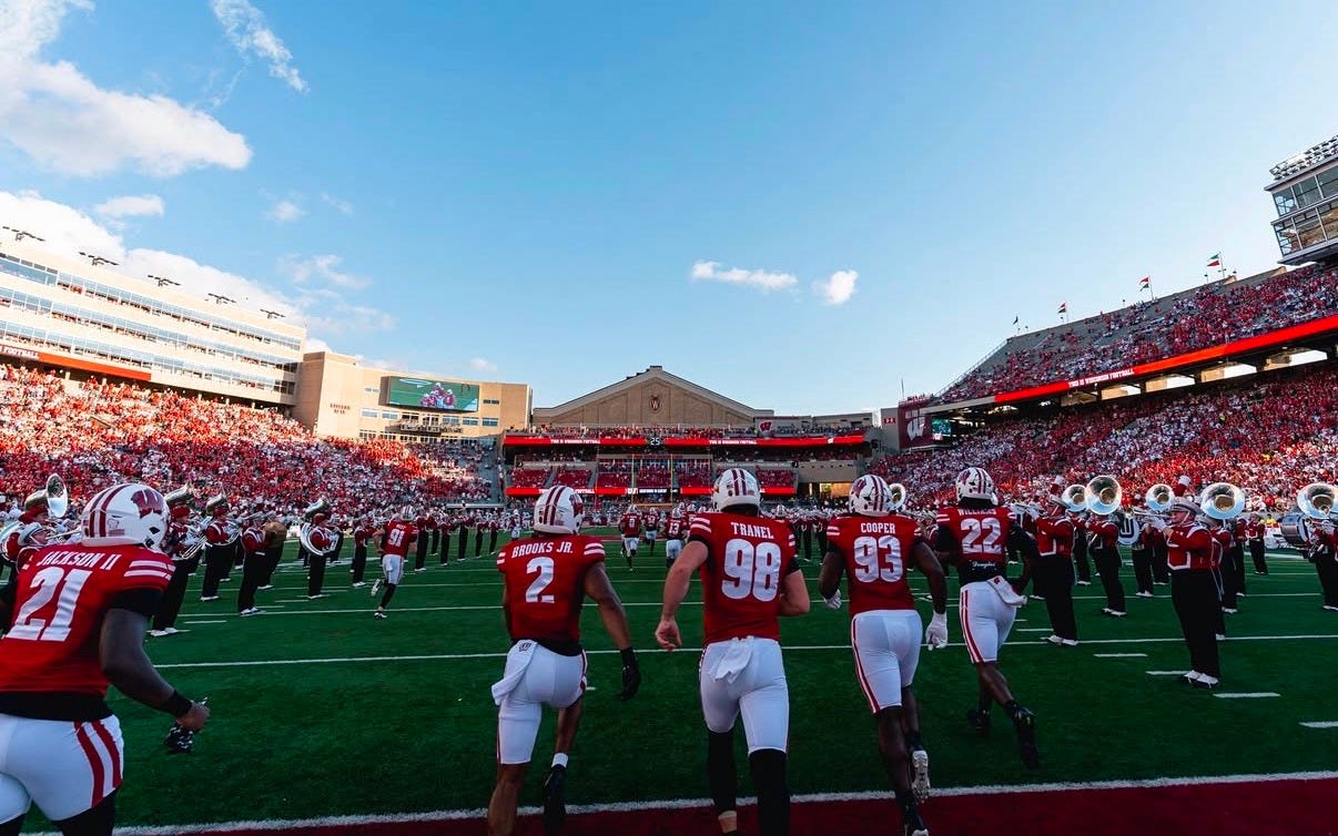 Wisconsin Badgers football players run onto the field at Camp Randall Stadium, surrounded by the marching band and fans in the stands.