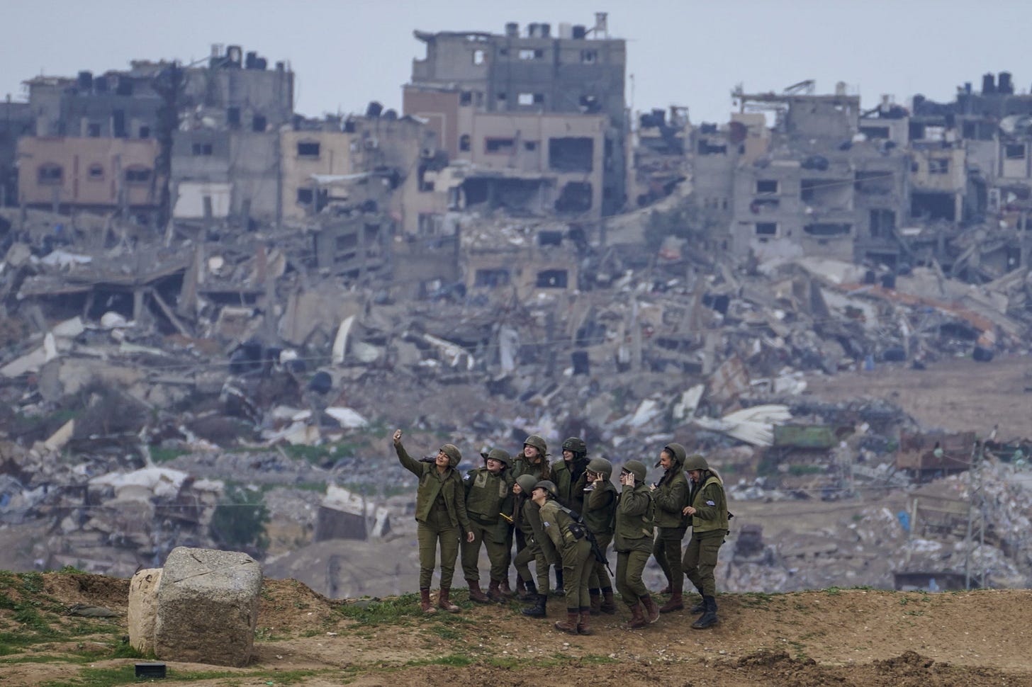 Ten smiling young women in Israeli military uniforms pose for a selfie on a hilltop overlooking destroyed buildings in Gaza.