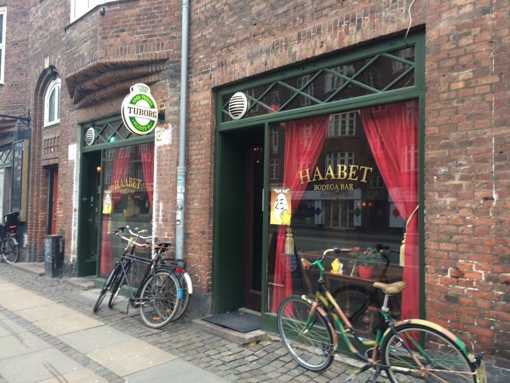 Set in a red brick building, two floor to ceiling windows make up Haabet Bodega - Tuborg sign lit up above the door and bikes parked outside