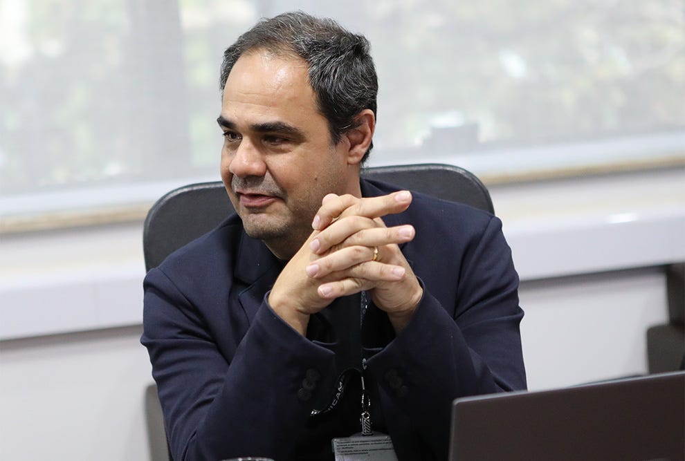 Pessoa sentada em cadeira de escritório, vestindo blazer azul-marinho sobre camisa preta, com crachá pendurado no pescoço, gesticula com ambas as mãos enquanto fala, tendo atrás de si um quadro branco e janela com vista desfocada de edifícios ao fundo. Pessoa sentada em cadeira de escritório, vestindo blazer azul-marinho sobre camisa preta, com crachá pendurado no pescoço, gesticula com ambas as mãos enquanto fala, tendo atrás de si um quadro branco e janela com vista desfocada de edifícios ao fundo.