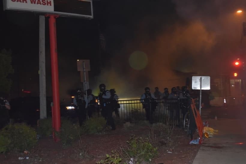A group of Minneapolis police officers in riot gear are in the distance, with weapons drawn. Clouds of tear gas are behind them. A group of Minneapolis police officers in riot gear are in the distance, with weapons drawn. Clouds of tear gas are behind them.