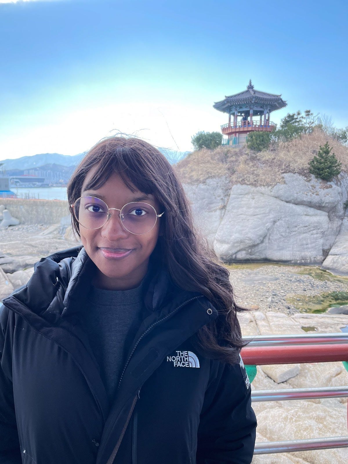 A headshot of Kat Lewis, a Black woman with long, dark hair and circular glasses, standing inside Yeonggeumjeong Sunrise Pavilion in Sokcho, South Korea with another pavilion behind her in the background. A headshot of Kat Lewis, a Black woman with long, dark hair and circular glasses, standing inside Yeonggeumjeong Sunrise Pavilion in Sokcho, South Korea with another pavilion behind her in the background.