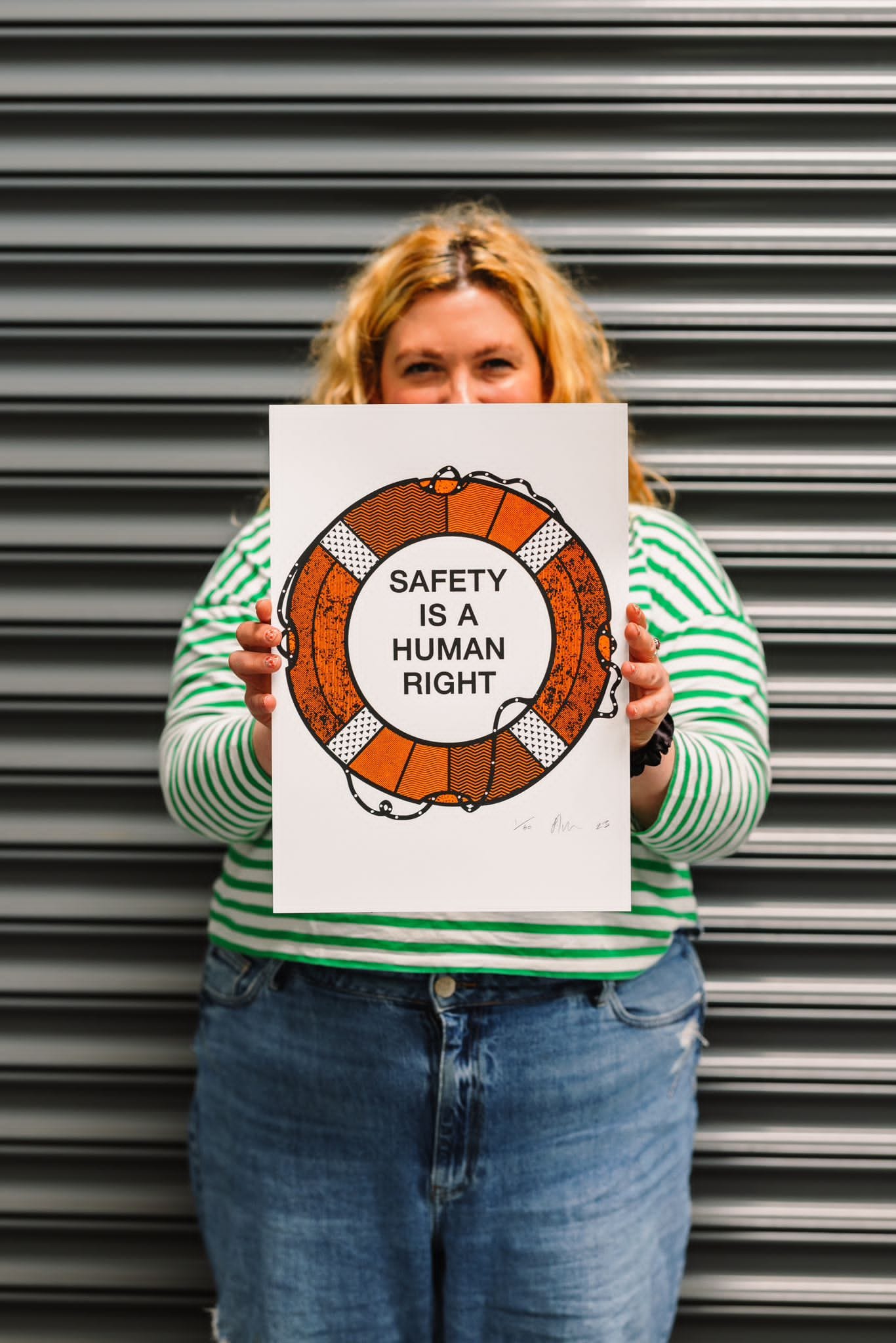 A woman stands in front of a closed shutter, she is holding up an artwork print of a dinghy, with text that reads 'safety is a human right', she is peering behind the print and looks happy.