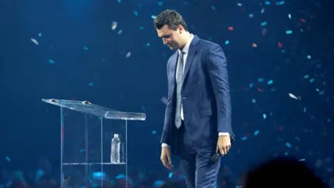Reuters Charlie Kirk stands in profile with confetti falling around him wearing a blue pinstripe suit on a darkened stage