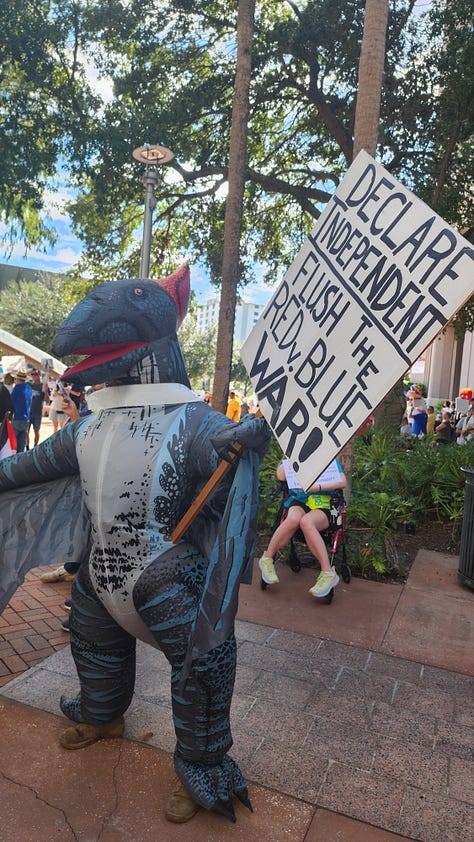 20251018_114518.jpg: A close-up of a hand-drawn protest sign reading "NO KINGS!!" and "NO WEENIES ALLOWED," featuring a crossed-out Burger King logo and a drawing of SpongeBob SquarePants.  20251018_113815.jpg: A wide shot of a large, sunny protest in a public plaza, showing crowds gathered around a modern arched sculpture and a water fountain.  20251018_113805.jpg: A crowd of protestors gathered on a brick plaza under a canopy of trees, with some people holding American flags and various signs.  20251018_110151 - Copy.jpg: A person in an inflatable blue pterodactyl costume stands at the protest, next to a person in a wheelchair holding a sign that reads, "DECLARE INDEPENDENT. FLUSH THE RED/BLUE WAR!"  20251018_113823.jpg: A wide view of the protest centered on a large, modern arched sculpture over a fountain, with a large crowd and many signs visible in the background.