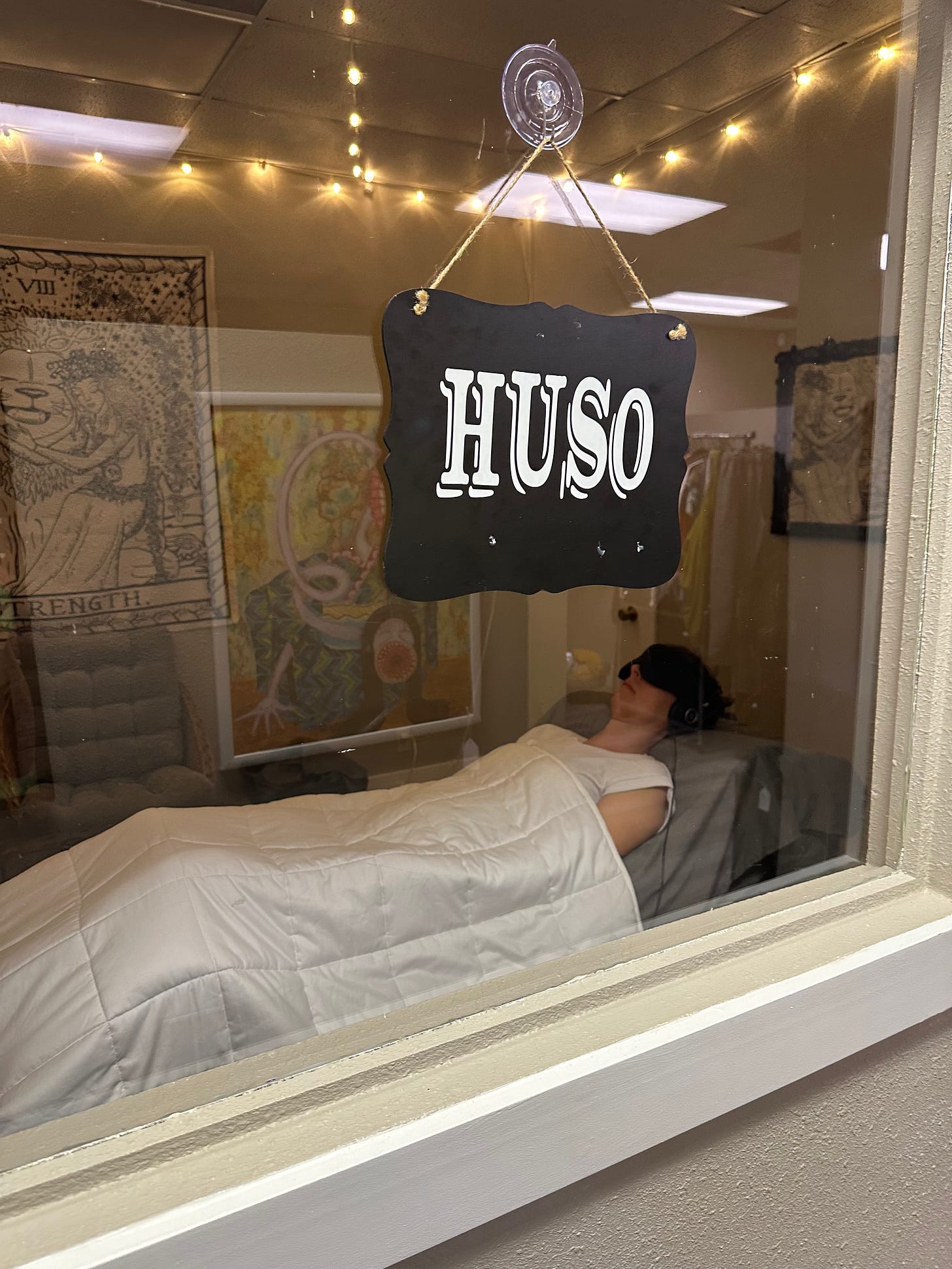Woman laying on a therapy table covered in a white blanket, with black headphones and a black eye mask. She's in a dimly lit therapy room with a sign that says "HUSO" on the window. Woman laying on a therapy table covered in a white blanket, with black headphones and a black eye mask. She's in a dimly lit therapy room with a sign that says "HUSO" on the window.