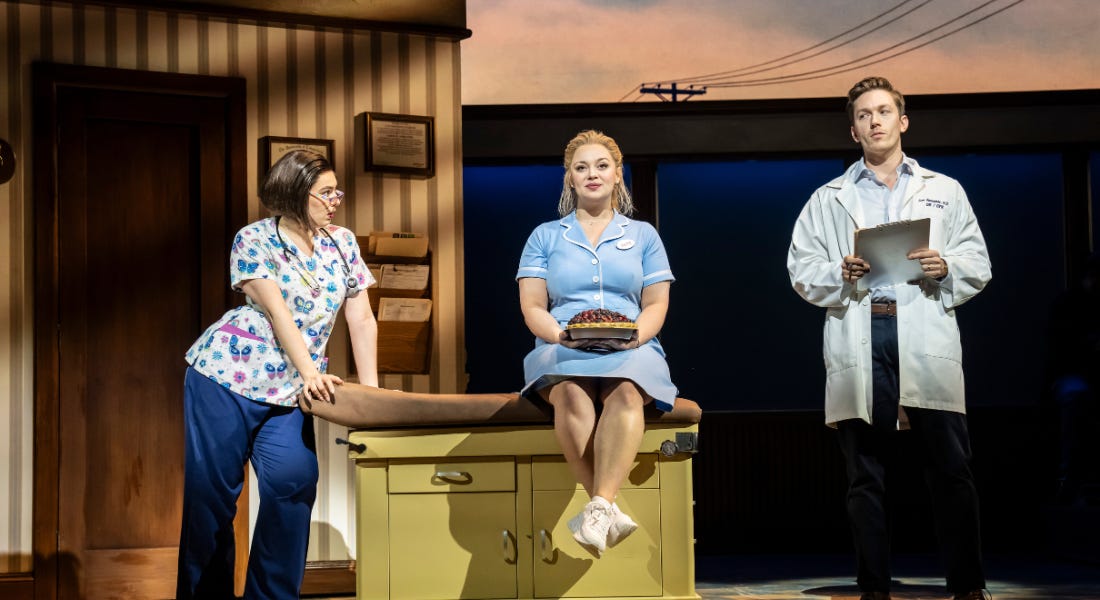 Three people on stage, with a woman in the middle sat on a yellow counter Three people on stage, with a woman in the middle sat on a yellow counter