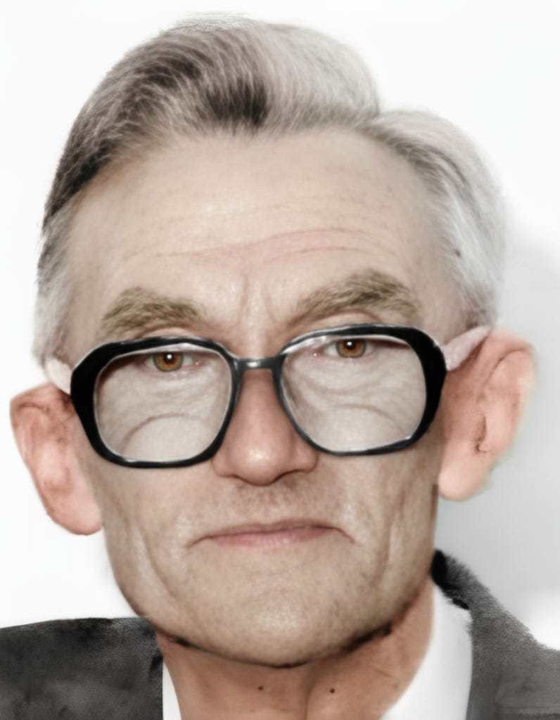 A close-up, colourised portrait of an older man with grey hair combed back from his forehead. He looks directly at the camera with a serious expression. He is wearing prominent, thick, dark-framed glasses in a horn-rimmed style. The shoulders of a dark suit jacket and the collar of a white shirt are visible at the bottom of the frame. The background is a stark, plain white.
