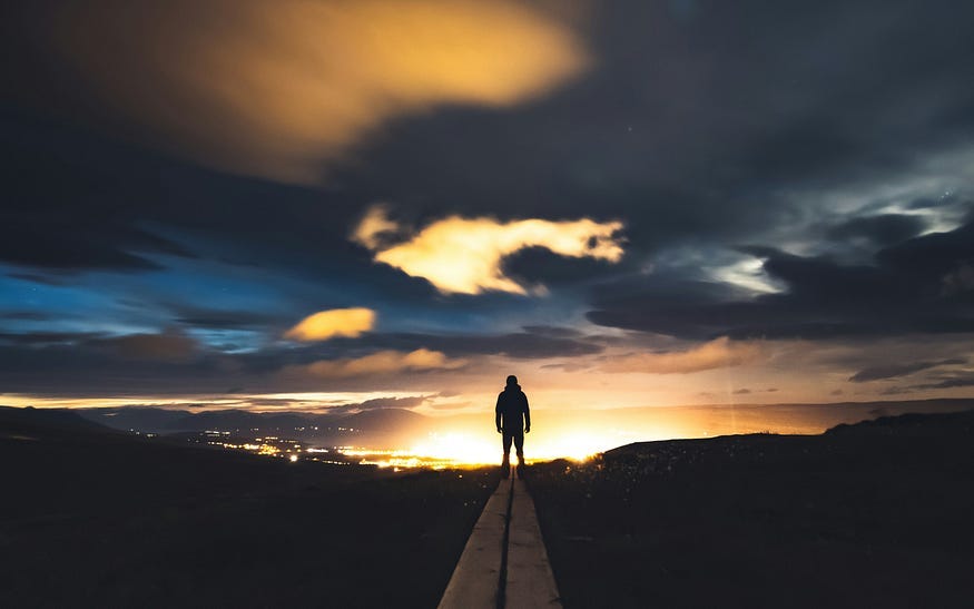 A figure stands in the middle of the road on the brow of a hill at night. Beyond, silhouetting them in the failing daylight, the glow of a city in the distance shines like a beacon.