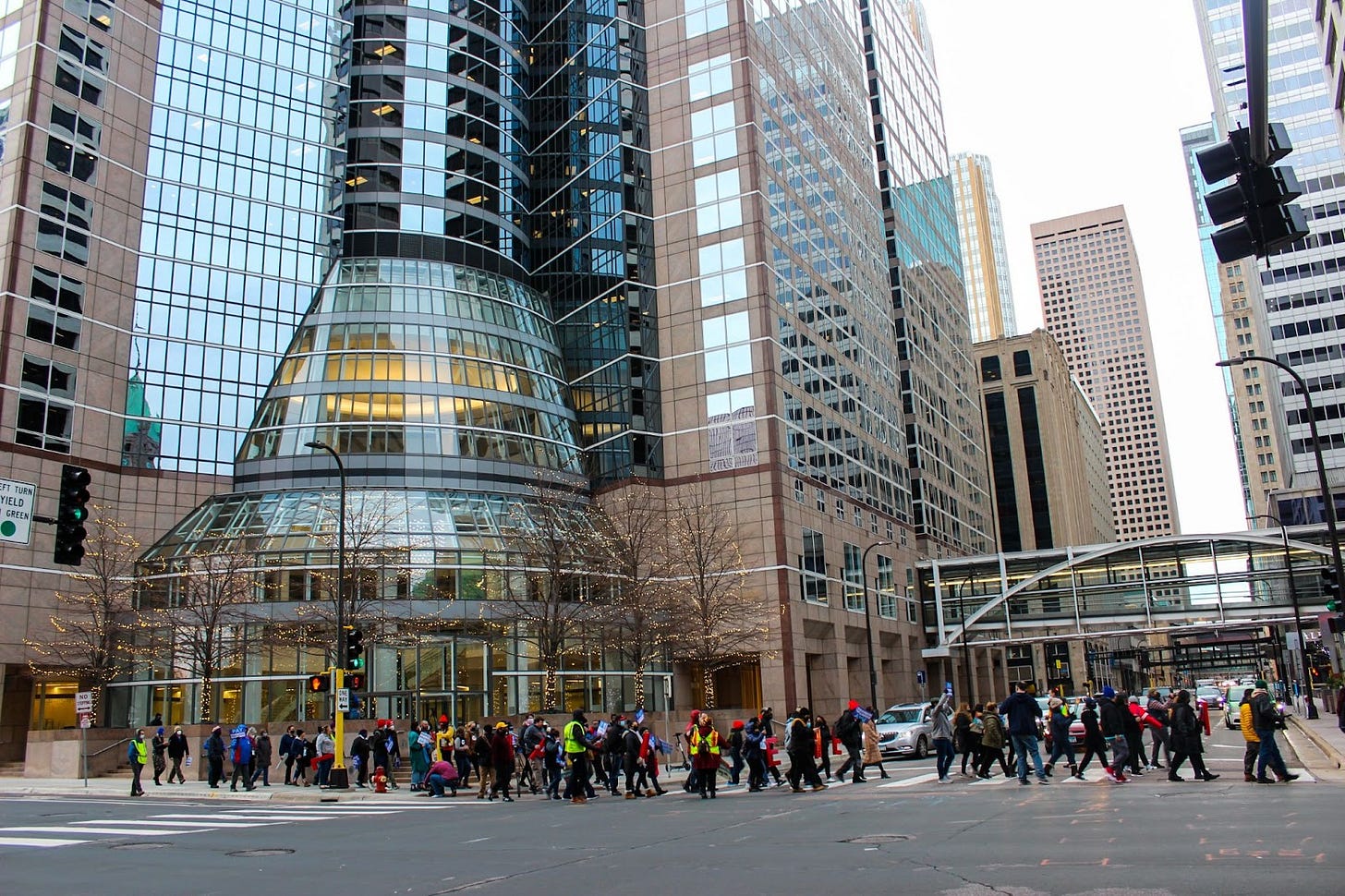 crowd of people march in front of tall glass building and tree with lights downtown