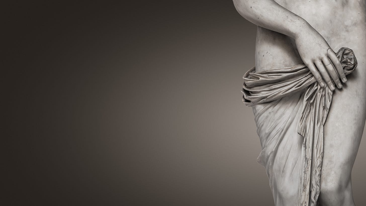 Close-up of a classical marble statue of a nude female figure, shown from the torso to upper thighs, with one hand resting near the hip and draped fabric partially covering the lower body against a neutral background.