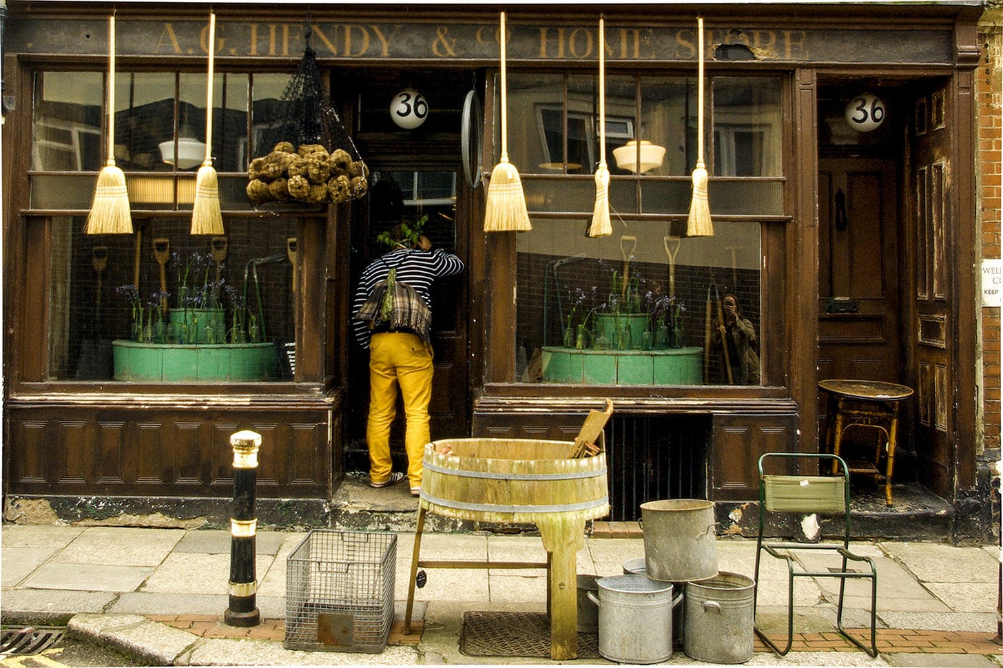 A vintage shopfront with brooms, garden tools and a person peering through the doorway. A vintage shopfront with brooms, garden tools and a person peering through the doorway.