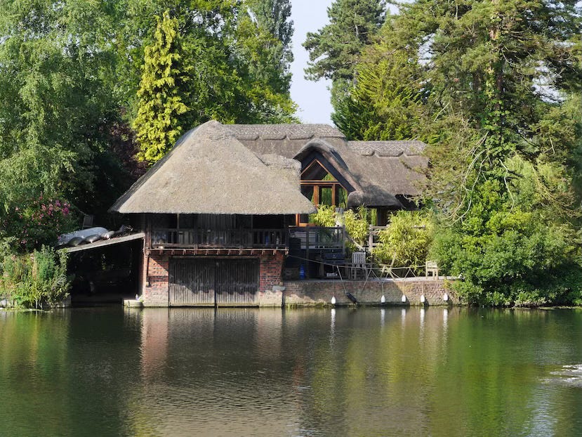  picturesque thatched boathouse sits nestled among lush trees on the edge of a calm river. The building features timber and brick construction with large glass windows and a deck area furnished with chairs and plants. A slipway and boat storage area are visible beneath the structure, while the surrounding greenery and reflections in the water add to the tranquil atmosphere.