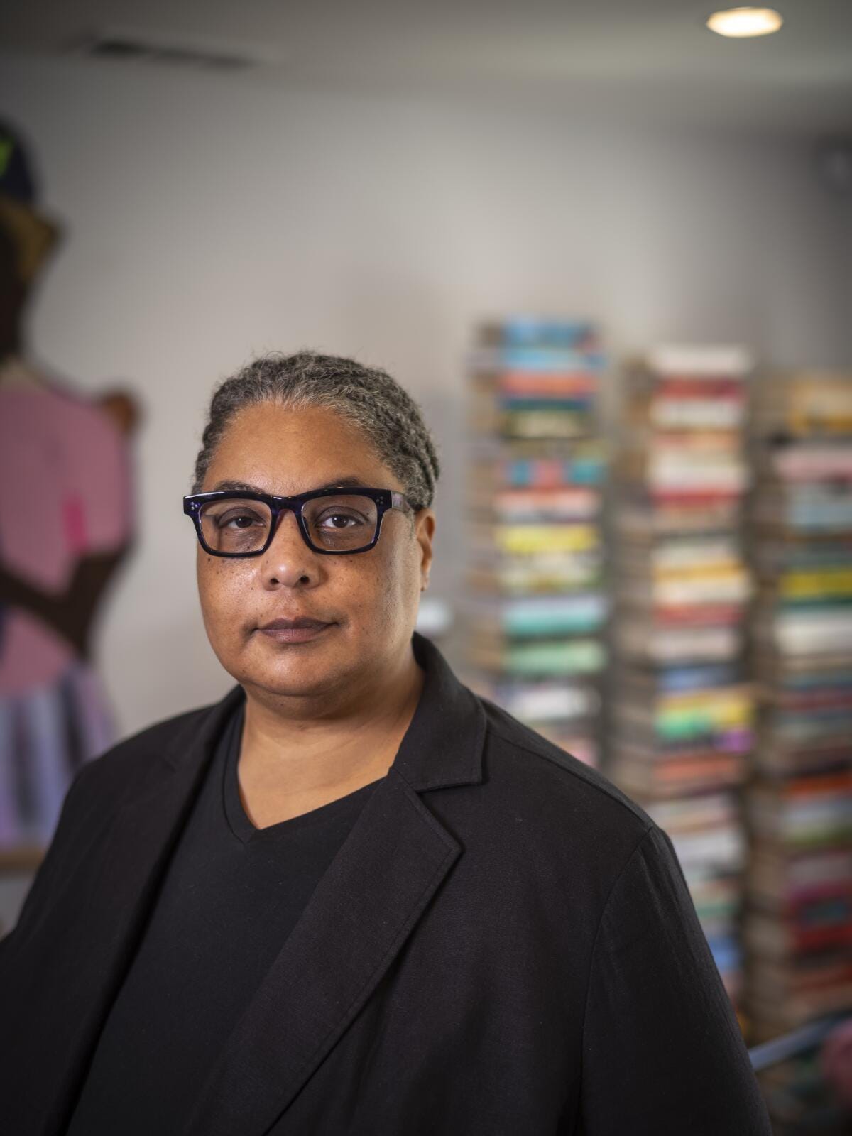Roxane Gay, wearing a black V-neck T-shirt, blazer and glasses, stands in front of three tall stacks of books. Roxane Gay, wearing a black V-neck T-shirt, blazer and glasses, stands in front of three tall stacks of books.