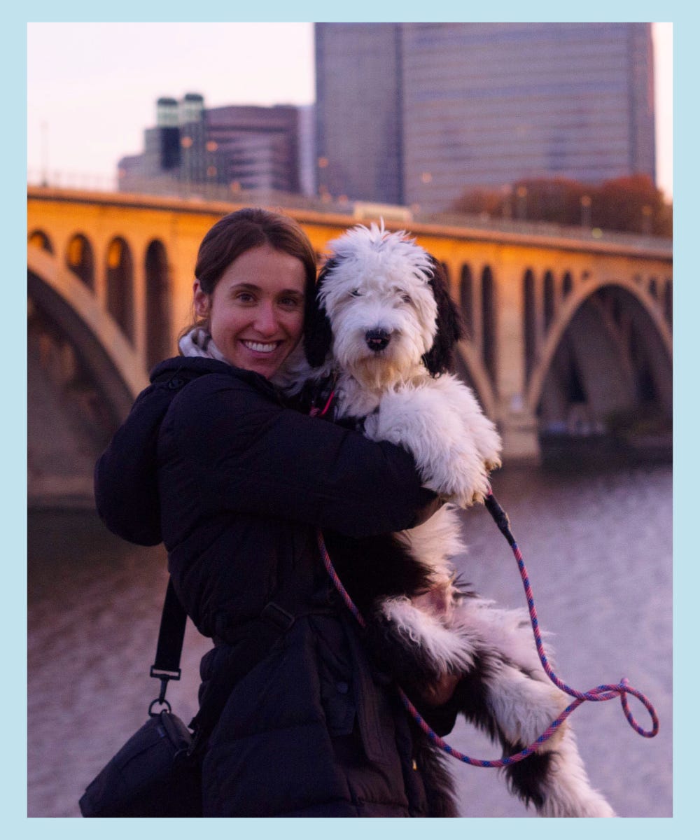 Christie holding Otis with his leash on in front of a bridge with buildings and water in the background
