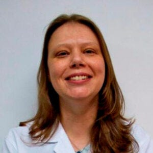 Mulher branca, de cabelo castanho comprido, usa um jaleco branco e sorri. No fundo, uma parede branca.