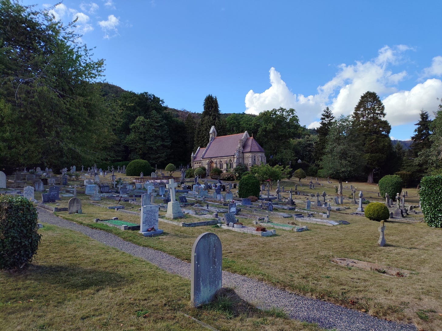 a graveyard with a small chapel in the background. The sky is blue and there is a wooded hill behind the graveyard.