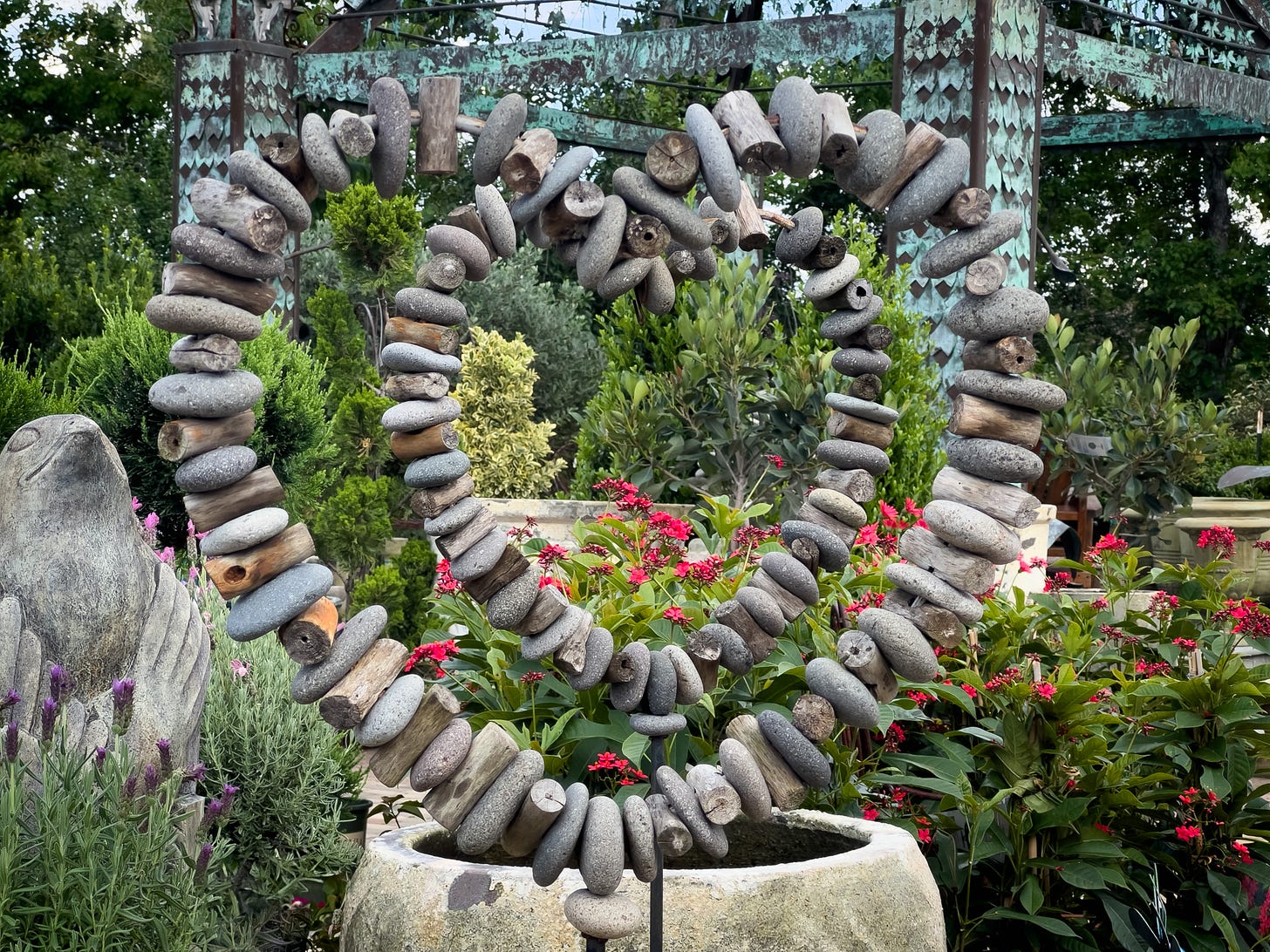 A heart-shaped sculpture made of stacked stones and driftwood, surrounded by vibrant flowers and lush greenery in a garden setting.