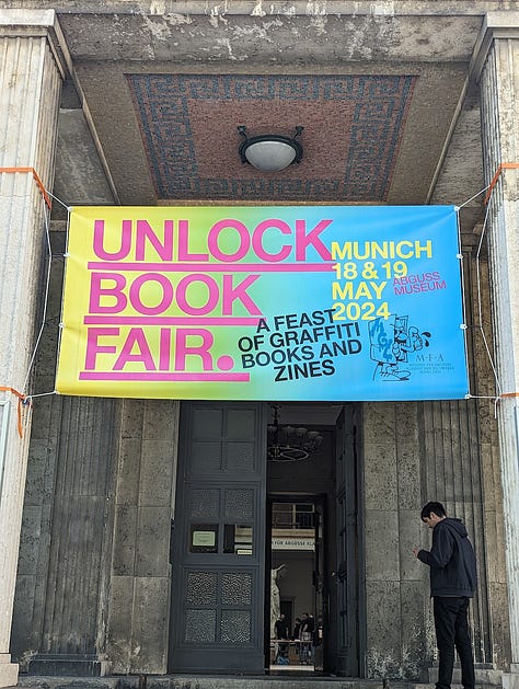 The first image shows a vibrant banner for the Unlock Book Fair in Munich, held on May 18-19, 2024, at the Abguss Museum. The banner is brightly colored with bold pink, yellow, and blue lettering, advertising the event as a feast of graffiti books and zines. The banner is hung above the entrance to a grand stone building with tall columns, as a person is seen checking their phone near the entrance. The second image captures a scene inside the book fair, where attendees browse through various graffiti books and zines at booths set up on long tables. In the foreground, a table prominently features Soi Books products, including zines, art books, and a selection of graffiti-related publications. The third image depicts two individuals at the book fair, one of whom is smiling and holding up a colorful graffiti art print by artist Tizer. The fourth image captures a lively moment in a traditional beer hall, where a group of people are enjoying drinks while a live band plays music in the background. One person is clapping along with the music, while others sit around the table chatting and drinking. The fifth image shows a classic Munich subway train arriving at a station, with its iconic blue and cream design. The train is captured just as it pulls into the platform, with a few passengers visible inside.