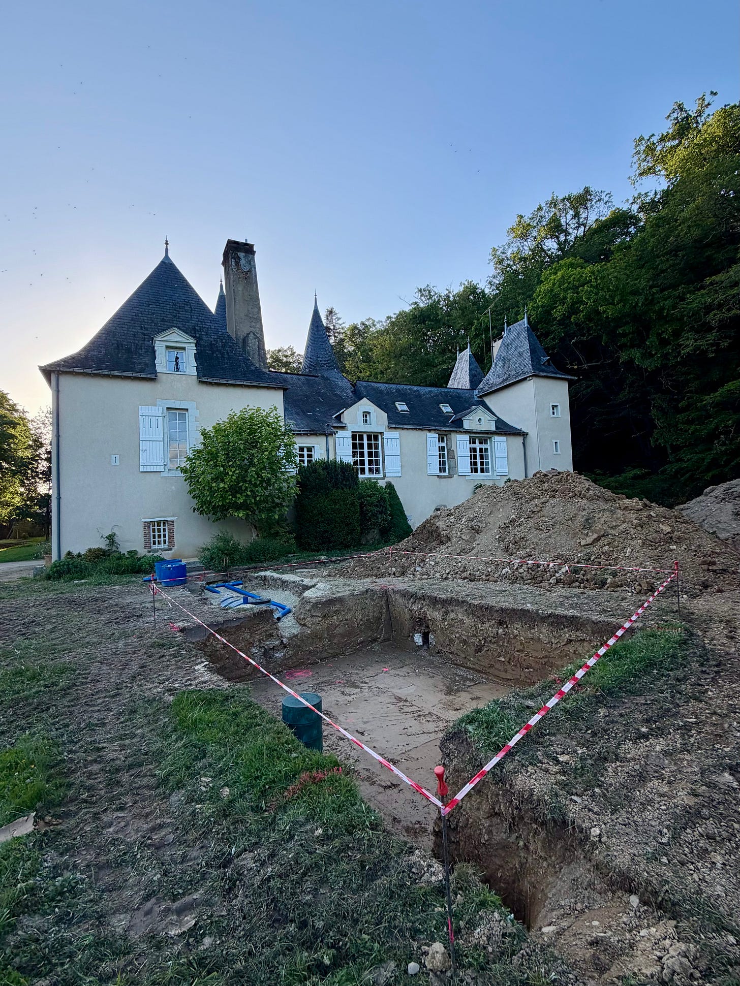 A deep rectangular excavation marked off with red and white tape in front of a cream château with white shutters and pointed slate turrets, with a tall mound of dirt beside it and blue pipes running into the trench. A deep rectangular excavation marked off with red and white tape in front of a cream château with white shutters and pointed slate turrets, with a tall mound of dirt beside it and blue pipes running into the trench.