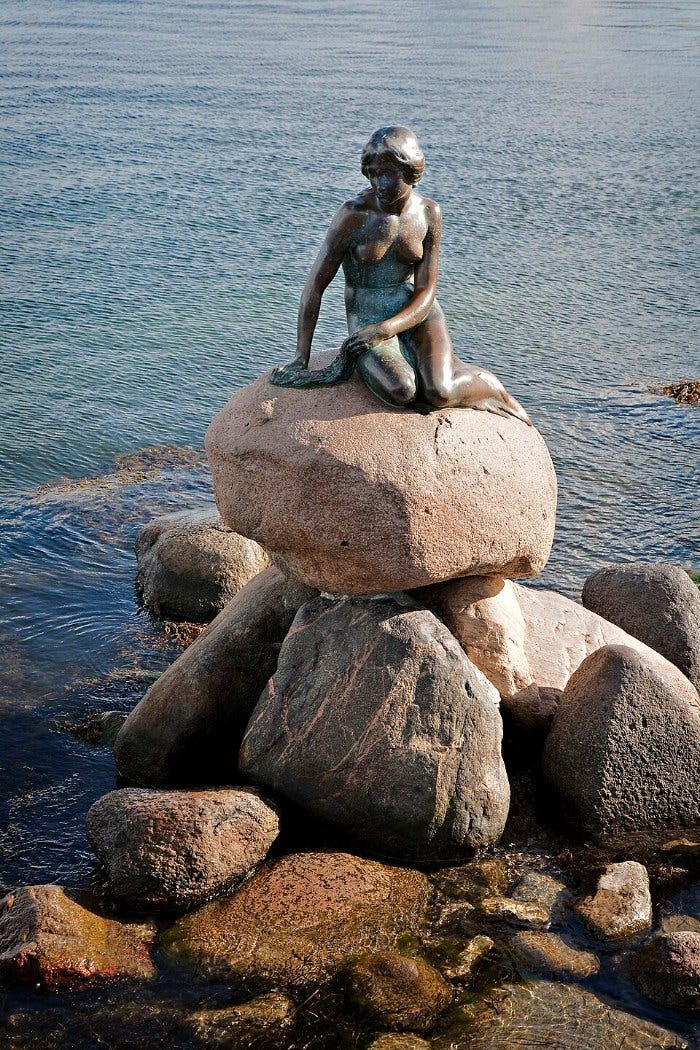 The small bronze Little Mermaid sitting on a pile of rounded boulders at the edge of Copenhagen harbor, her fish tail curled beneath her The small bronze Little Mermaid sitting on a pile of rounded boulders at the edge of Copenhagen harbor, her fish tail curled beneath her