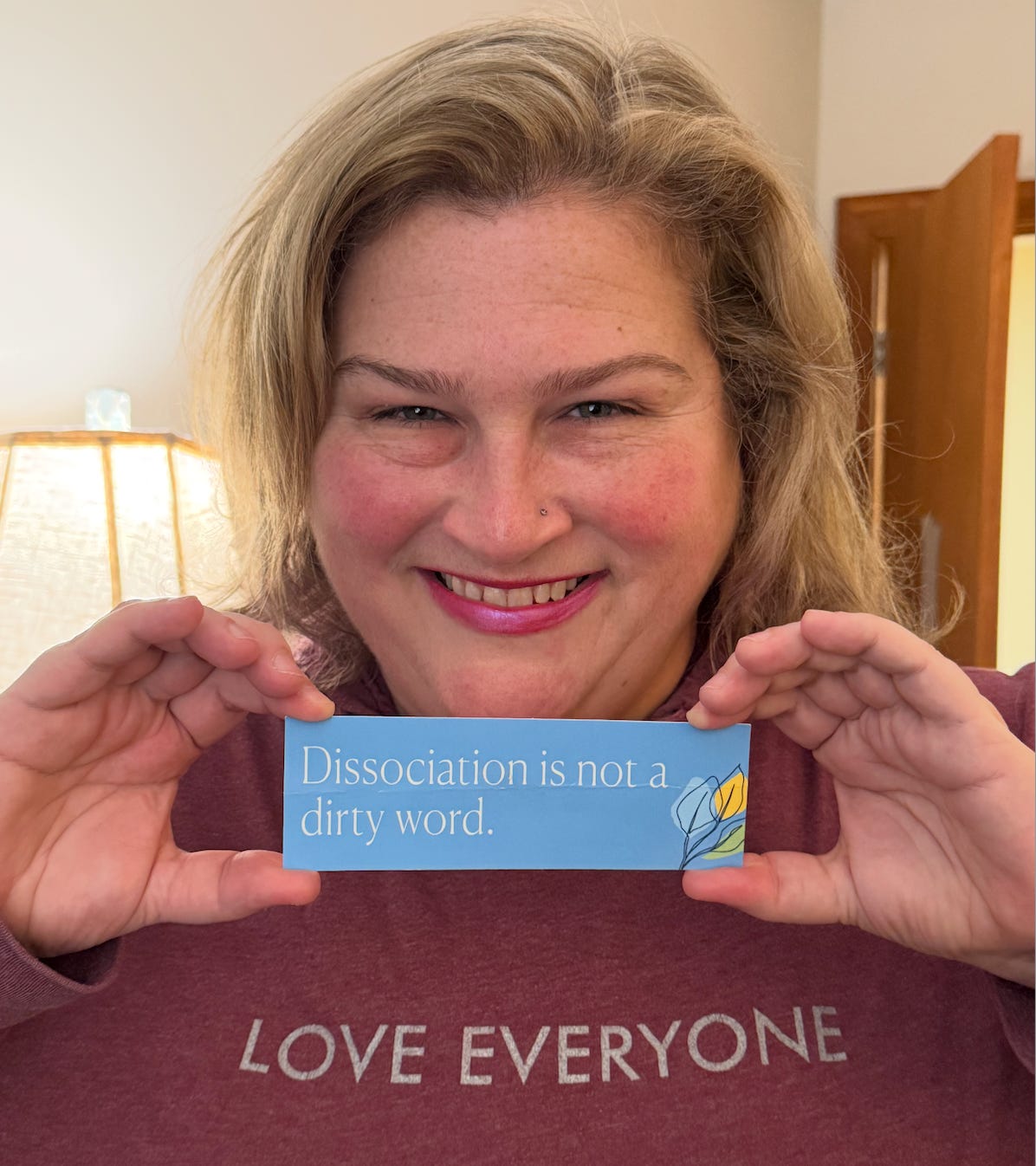 Jamie a blonde woman smiles holding the bookmark "Dissociation is not a dirty word"