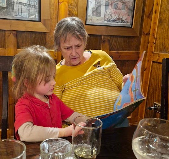 a picture of a 4 year old with blond hair and an woman in her late 60s reading book at a table full of empty galsses a picture of a 4 year old with blond hair and an woman in her late 60s reading book at a table full of empty galsses
