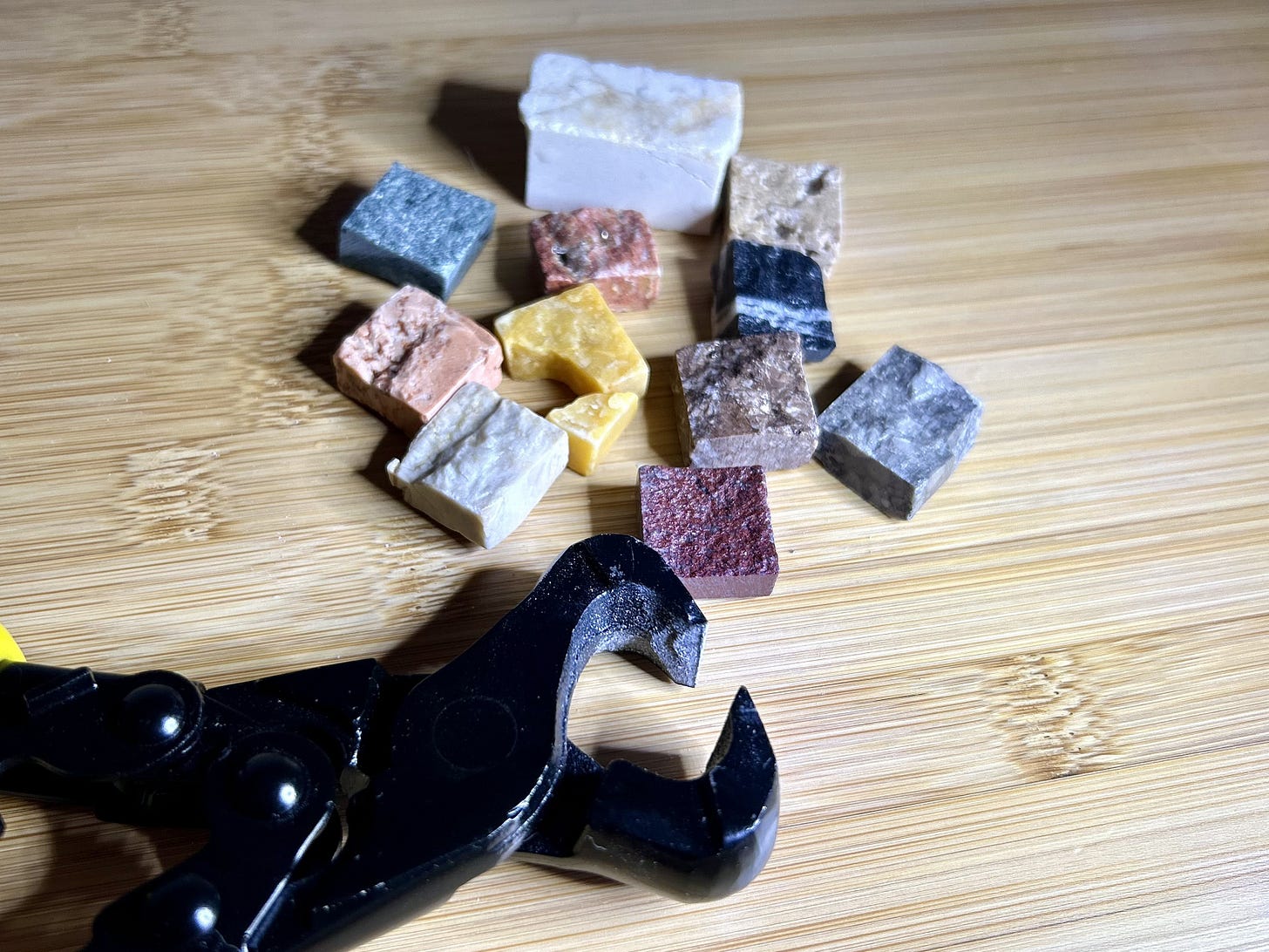 in the foreground a pair of black metal tile nippers with stone mosaic tesserae in various colours behind them on a bamboo tabletop.