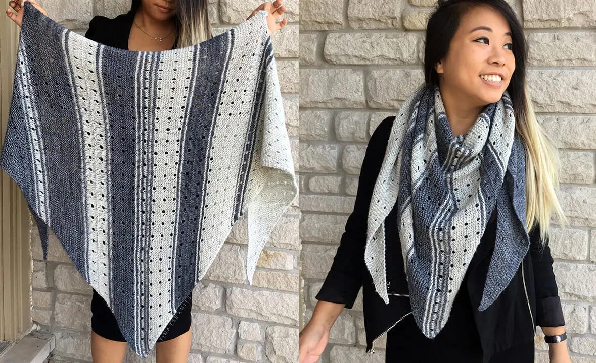 woman stands in front of a white brick wall wearing a bluish-gray and white knitted triangle shawl.