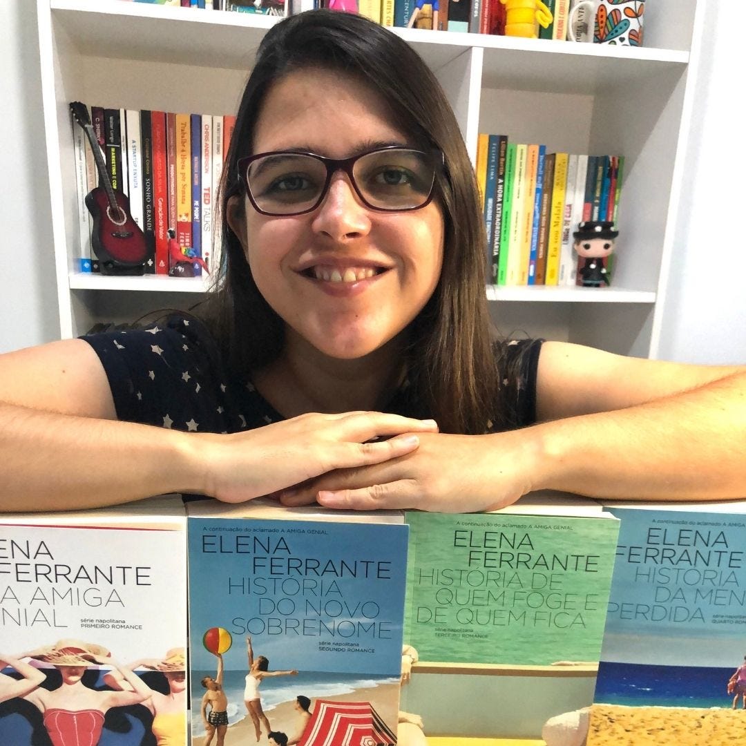 Foto de Cíntia, cabelos longos e lisos, de óculos, com estante branca de livros atrás e braços apoiados nos 4 livros da tetralogia da Amiga Genial, todos de pé, um ao lado do outro.