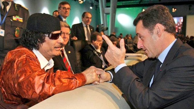 Muammar Gaddafi wearing a black cap and sunglasses, dressed in an orange jacket, shaking hands with Nicolas Sarkozy, who is in a dark suit. They are at an event with other people in suits and uniforms visible in the background, standing behind a barrier.