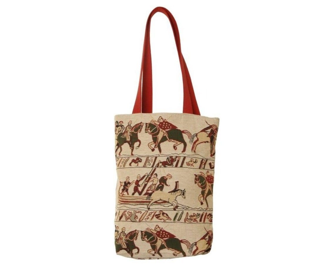 May include: A beige tote bag with red handles. The bag is decorated with a tapestry-like design featuring a scene of people riding horses. The scene is in shades of brown, green, and red.