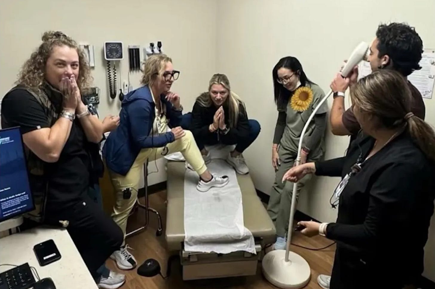 Six nurses stand gathered around a medical table, posing together. On the table, a sanitary sheet is spread out with a visible medical discharge sample.