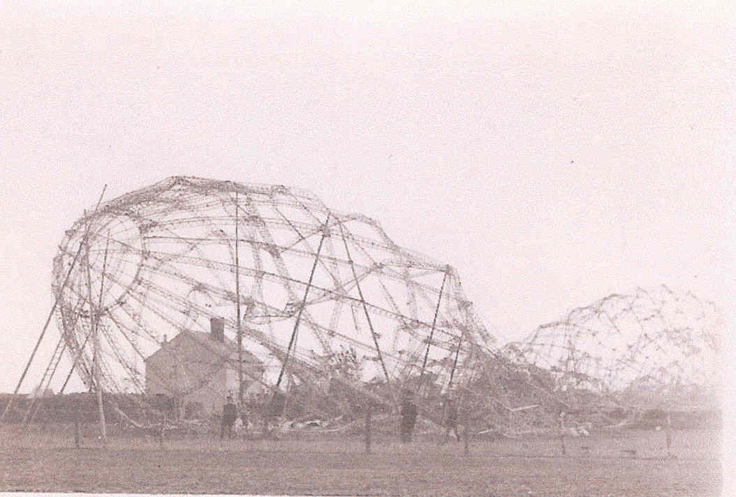 A photograph of the L33 wreck. The metal framing is all that remains. In the background a two storey house is dwarfed by the wreckage, which is about 3 times the size. In the foreground three people can just about be seen.