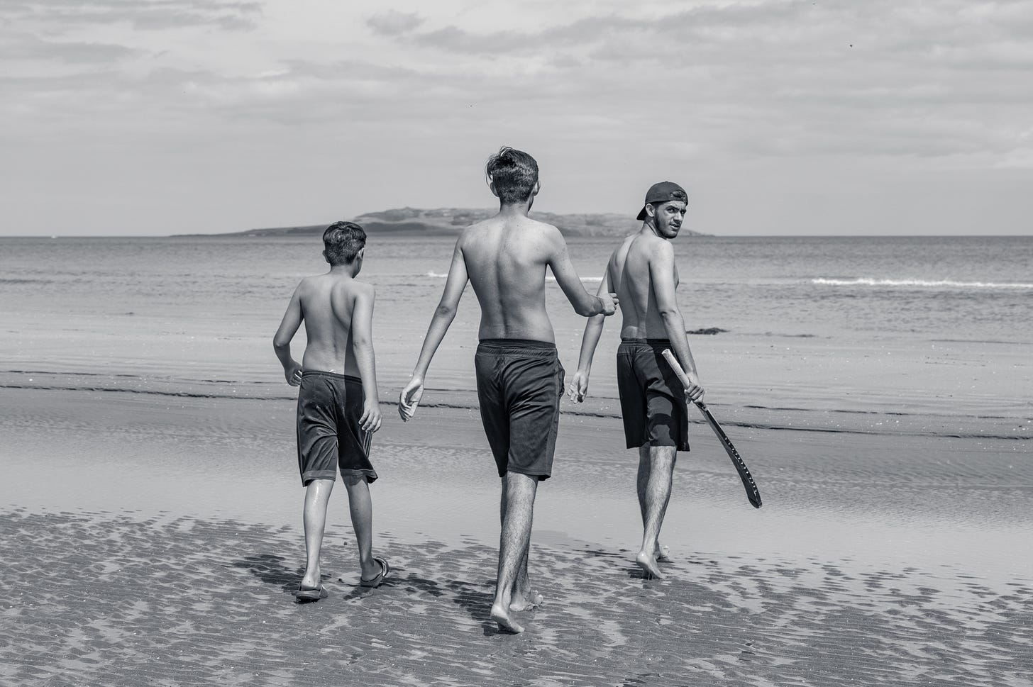 Three young lads in swim shorts walking away from the camera down a wide empty strand towards the sea, in black and white, a headland in the distance. Three young lads in swim shorts walking away from the camera down a wide empty strand towards the sea, in black and white, a headland in the distance.