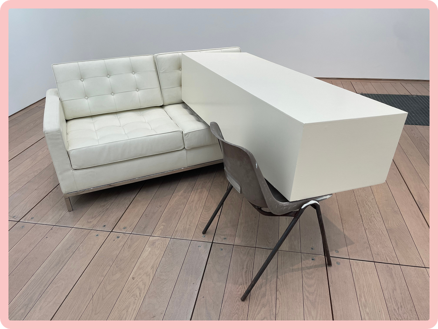 A photo of an installation in a white-walled gallery space, in which a long ivory-coloured box (resembling a coffin), is suspended between a white leather sofa and a grey institutional chair. A photo of an installation in a white-walled gallery space, in which a long ivory-coloured box (resembling a coffin), is suspended between a white leather sofa and a grey institutional chair.