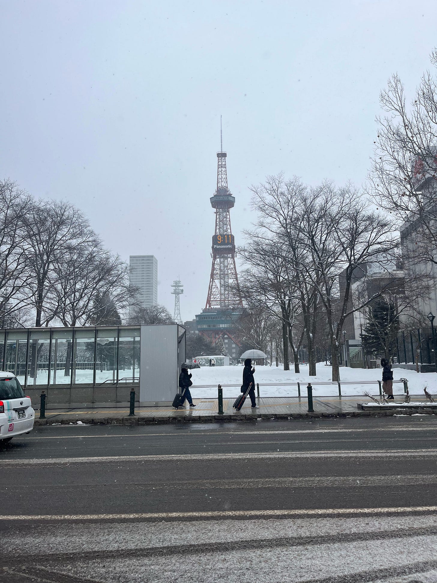 Sapporo TV Tower with the time 9:11 AM on it. Bare trees surround the flat park. There are tire marks with snow on the pavement in front and a person walking by with their transparent plastic umbrella.