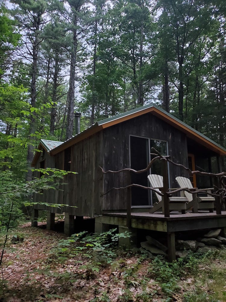 A rustic cabin of dark wood with two Adirondack chairs on the porch, surrounded by green foliage in the New England woods. 