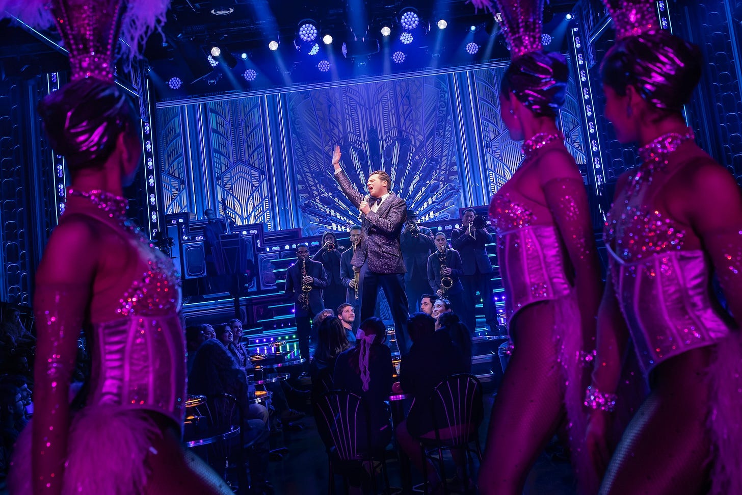 Groff sings into a handheld mic again, flanked by showgirls again Groff sings into a handheld mic again, flanked by showgirls again
