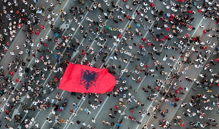 An aerial view of people in the street. Multiple people hold up a big Albanian flag. An aerial view of people in the street. Multiple people hold up a big Albanian flag.