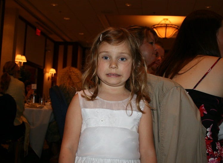 A little girl, five or six years old, all dressed up in a white dress for a wedding reception, gives a forced, nervous smile to the photographer. Her arms are tight at her side and her smile is decidedly not spontaneous or happy.