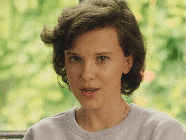 Giff da atriz Millie Bobby Brown, com cabelos castanhos curtos acima dos ombros, colocando o dedo indicador na frente dos lábios entreabertos, como quem diz "shhh".