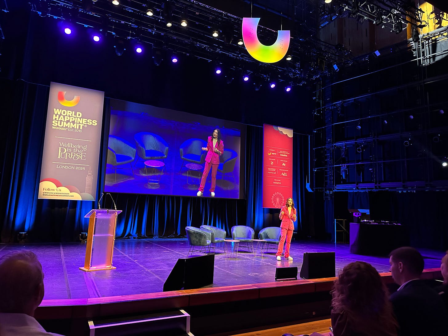 Karen Guggenheim wears a pink suit and stands on a stage in front of a sign reading World Happiness Summit, WOHASU Est 2016. Wellbeing is the Purpose London 2014. 