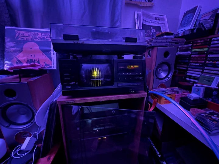 L to r: a photograph of a 100-disc CD changer stacked  and surrounded by newspapers, records, and other ephemera. the lighting of the photo is blue; a corner of a living room in which a lamp stands. 3 windows with the blinds raised can be seen. a small table with speakers and plants is also in the room.