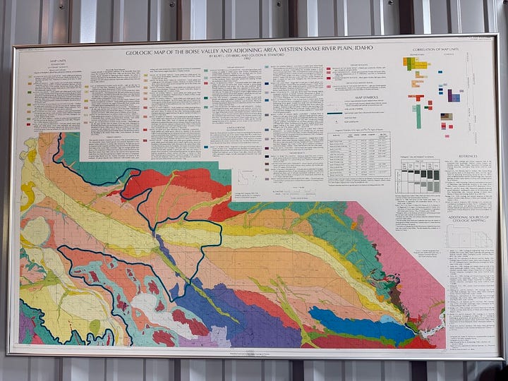 A gallery from Scoria Vineyards in Caldwell, Idaho: a rusted metal 'S' sign on the exterior of the tasting room barn overlooking the vineyard, and a framed geologic map of the Boise Valley and western Snake River Plain on display inside.