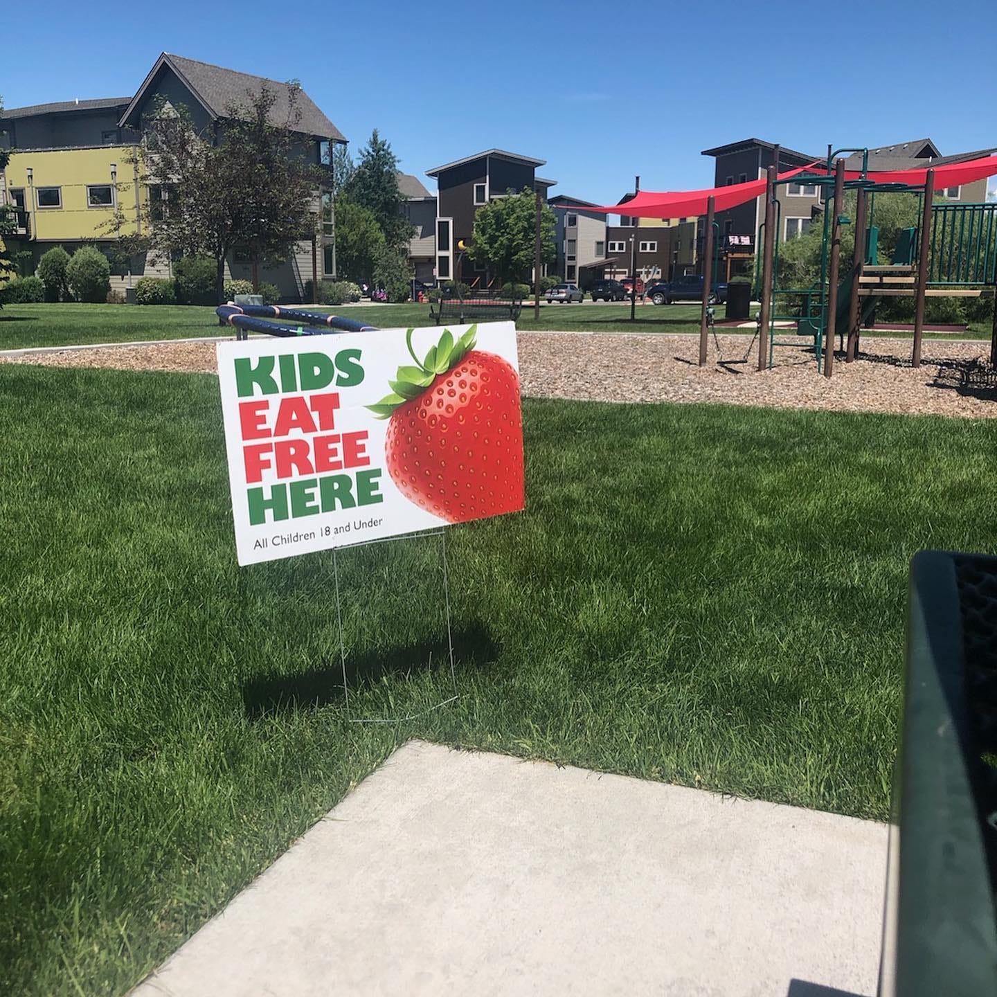 A park with a playground and green grass. There is a sign in the grass that says "Kids Eat Free Here"