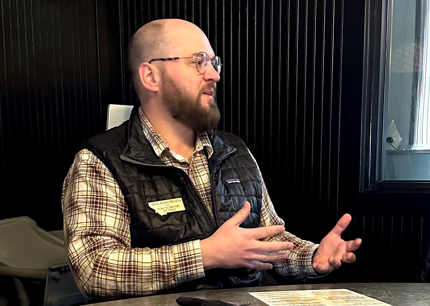 man with shaved head, beard and glasses speaking inside in black vest and flannel, seated