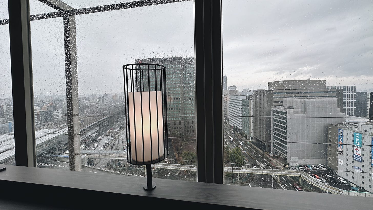 A single lamp glowing in front of windows overlooking Osaka buildings through windows covered in rain.