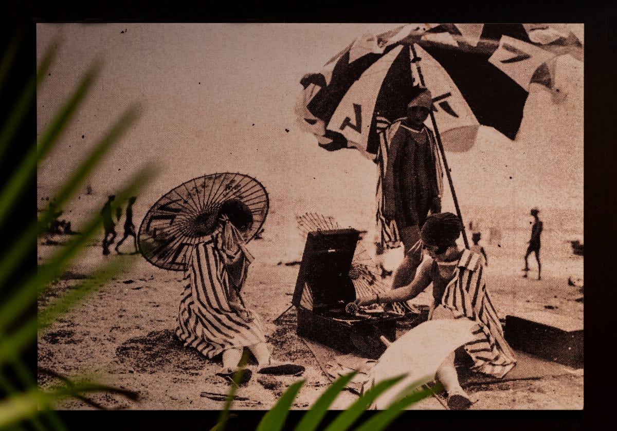 A grainy, sepia photo shows three women at the beach, wrapped in striped towels and listening to a portable gramophone player